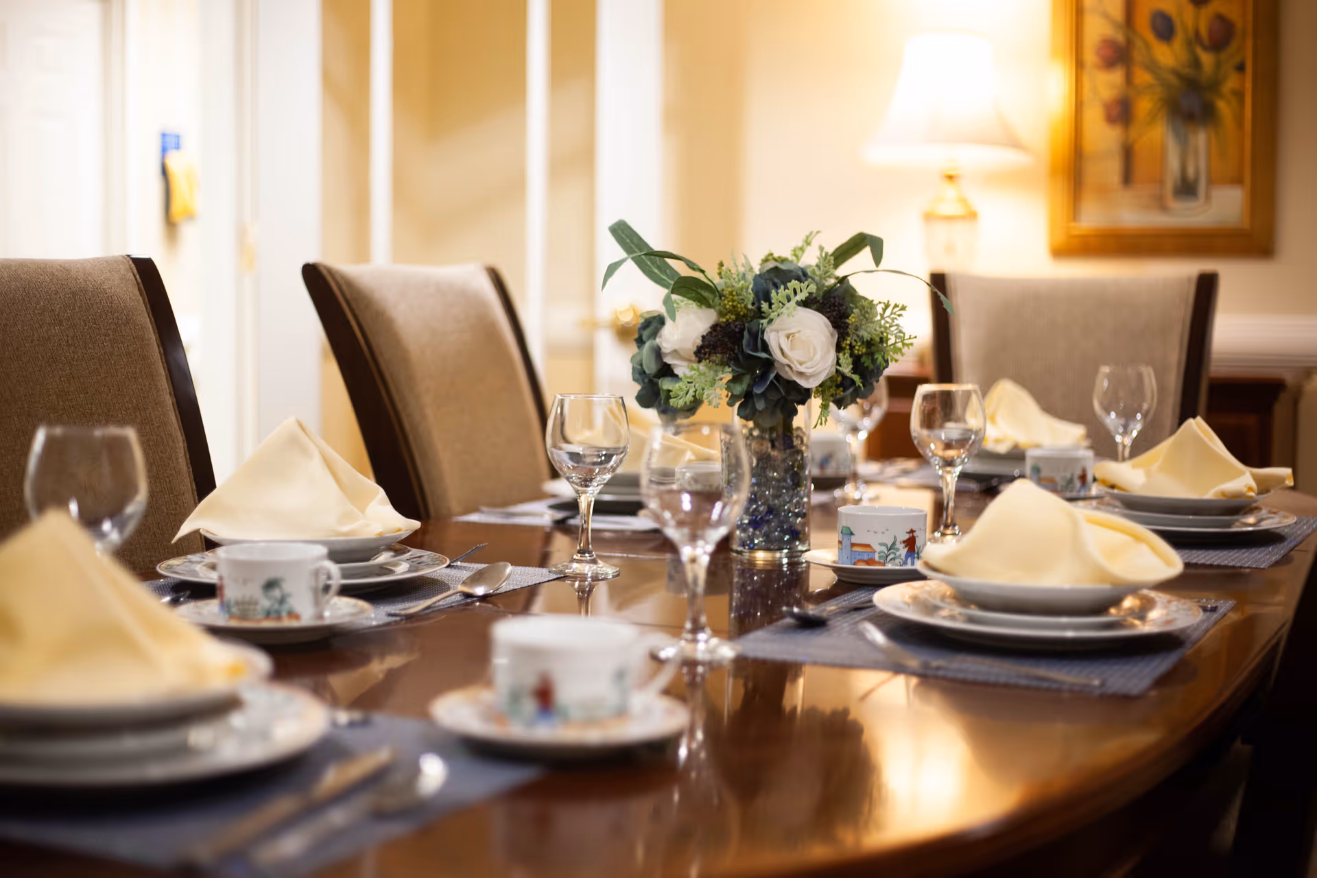 A formal dining table set with plates, cups, silverware, wine glasses, and folded yellow napkins. A floral centerpiece with white and green flowers is placed in the middle of the table. The room has beige walls, a lamp, and a framed painting in the background.