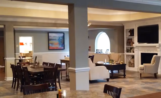 Interior view of a senior living facility common area with a dining section featuring multiple wooden tables and chairs, and a living room area with beige armchairs, a coffee table, a fireplace, and a mounted flat-screen TV. The room has neutral-colored walls, a large arched window, and decorative artwork on the walls.