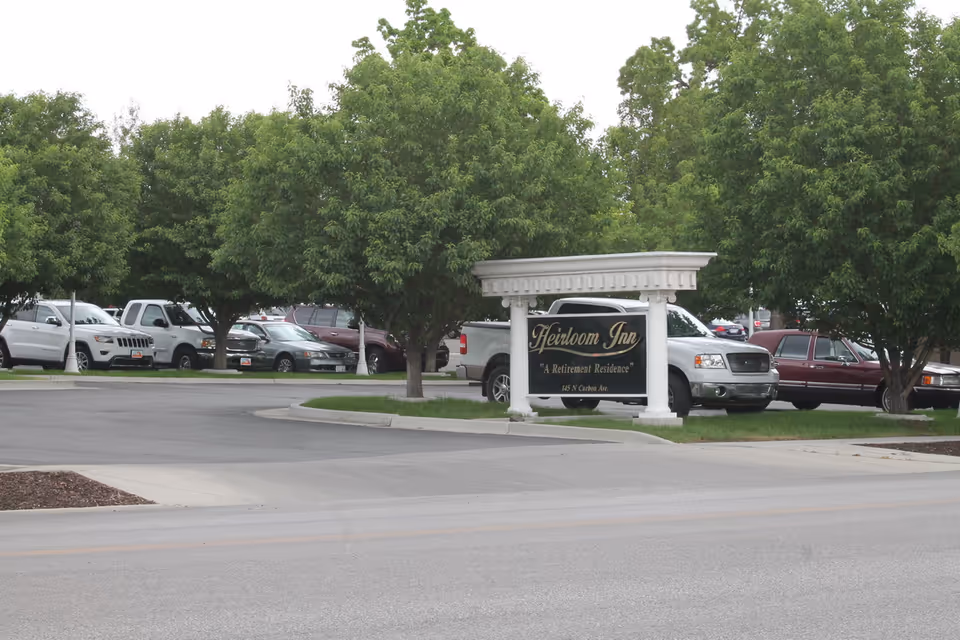 Parking lot area with several parked cars and green leafy trees surrounding a white sign that reads 'Heirloom Inn, A Retirement Residence, 145 N Carbon Ave.'