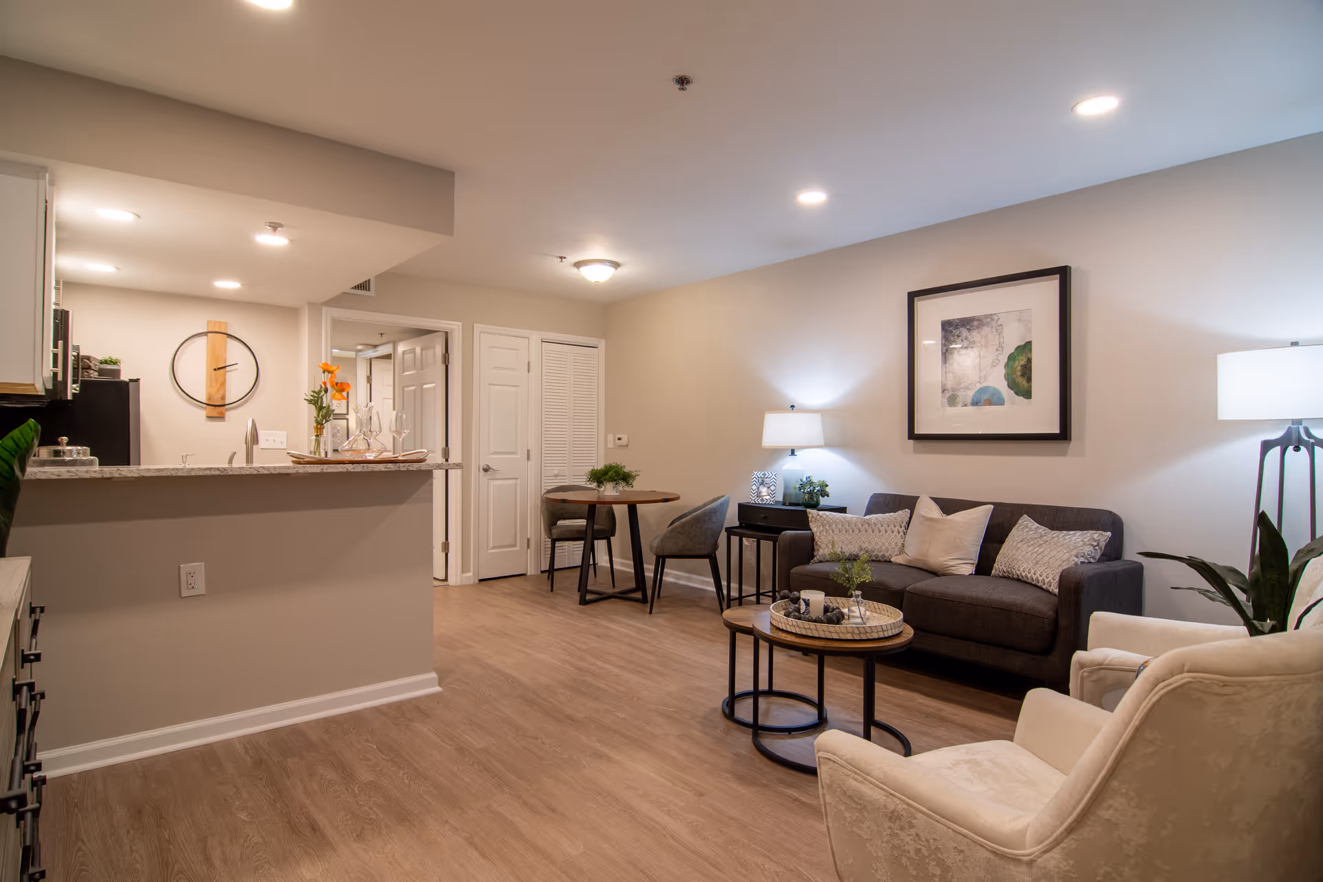 A cozy living room area in a retirement community featuring a dark gray sofa with multiple pillows, a round wooden coffee table with decorative items, a beige armchair, a floor lamp, and a side table with a lamp and plant. The room has light-colored walls and wood flooring. In the background, there is a small dining table with two chairs and a kitchen counter with a granite countertop and a wall clock above the sink.