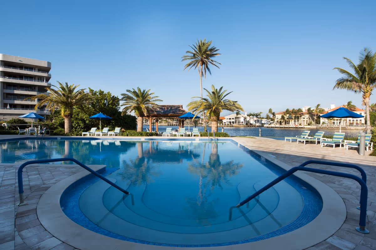 Outdoor swimming pool with clear blue water surrounded by palm trees, lounge chairs with blue umbrellas, and a waterfront view with buildings in the background under a clear blue sky.