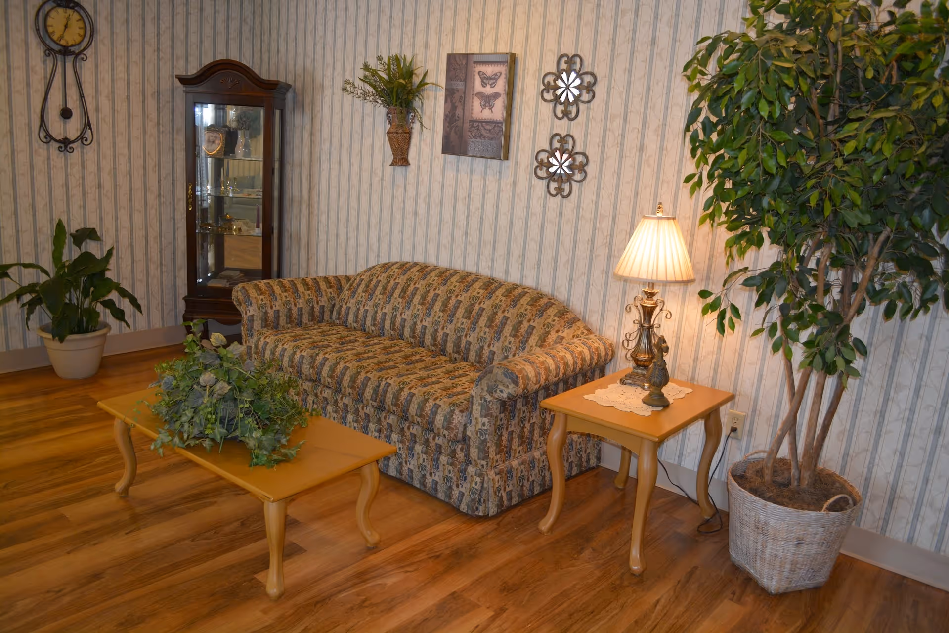 A cozy living room area with a patterned sofa, a wooden coffee table with a green plant centerpiece, a wooden side table with a decorative lamp and small figurine, a large potted plant in a wicker basket, a glass display cabinet, and wall decorations including a clock, a butterfly picture, and two floral metal wall hangings.