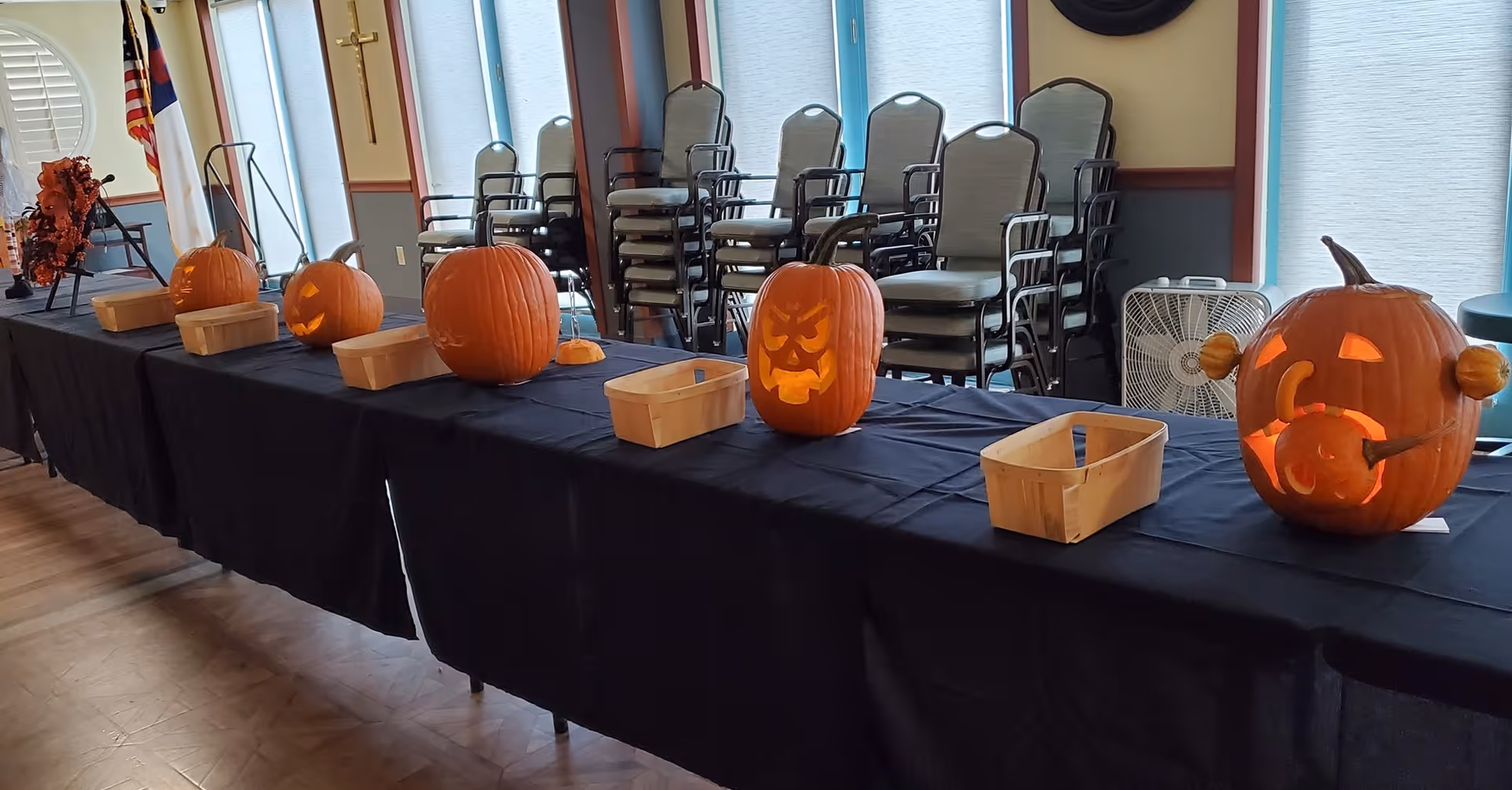 A long table covered with a black tablecloth displaying several carved pumpkins with various facial expressions. Behind the table, there are stacked chairs and a fan near windows with blinds. The setting appears to be an indoor room with wooden flooring and flags in the background.