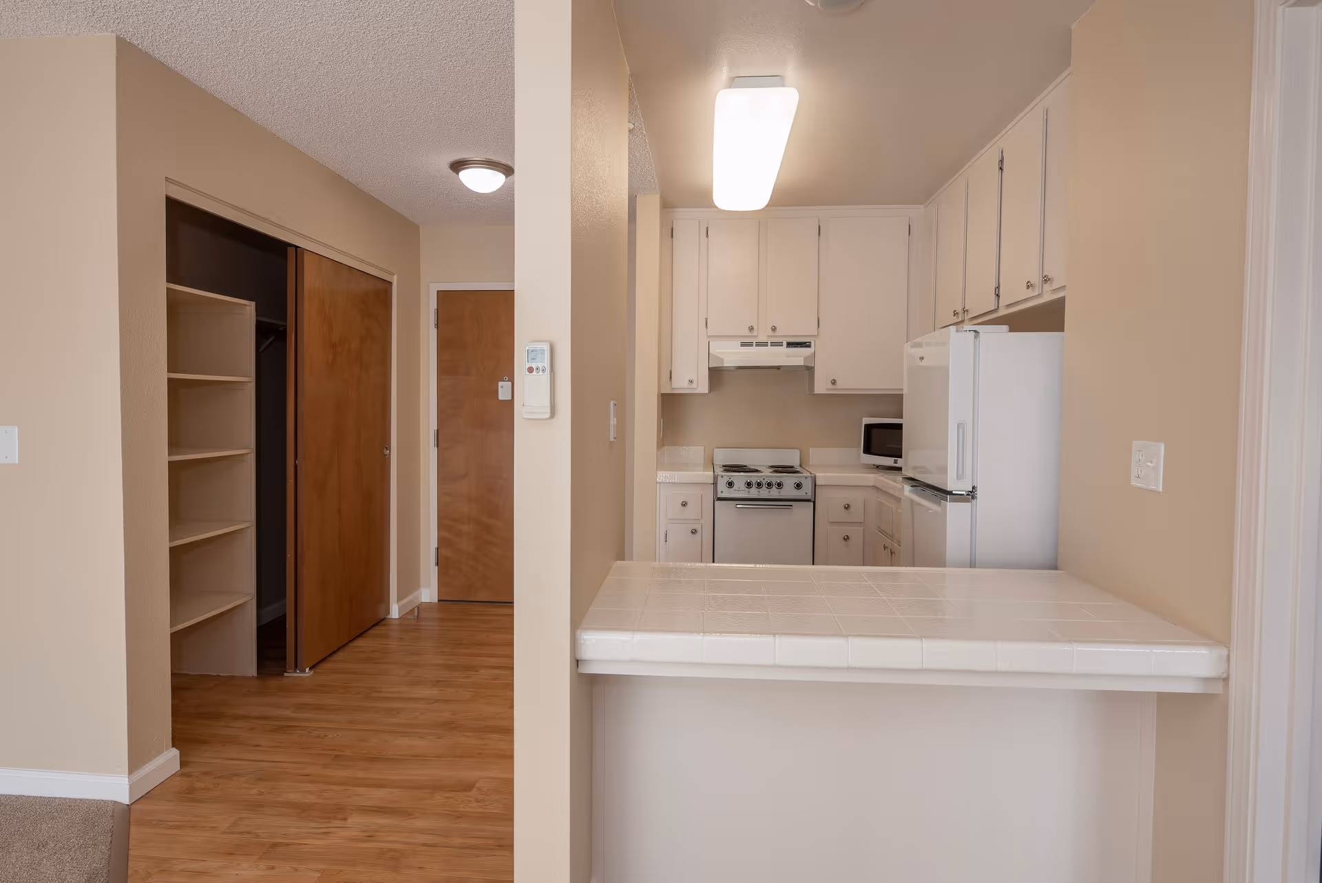 Interior view of a senior living facility kitchen with white cabinets, a white refrigerator, a small stove, and a microwave. The kitchen has a tiled countertop bar area facing an open space with wooden flooring and a closet with sliding wooden doors.