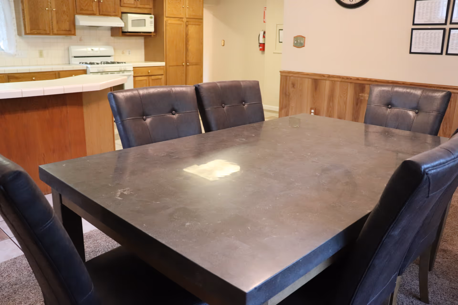 Large dark dining table surrounded by six leather-upholstered chairs in a dining area with a view of the kitchen.