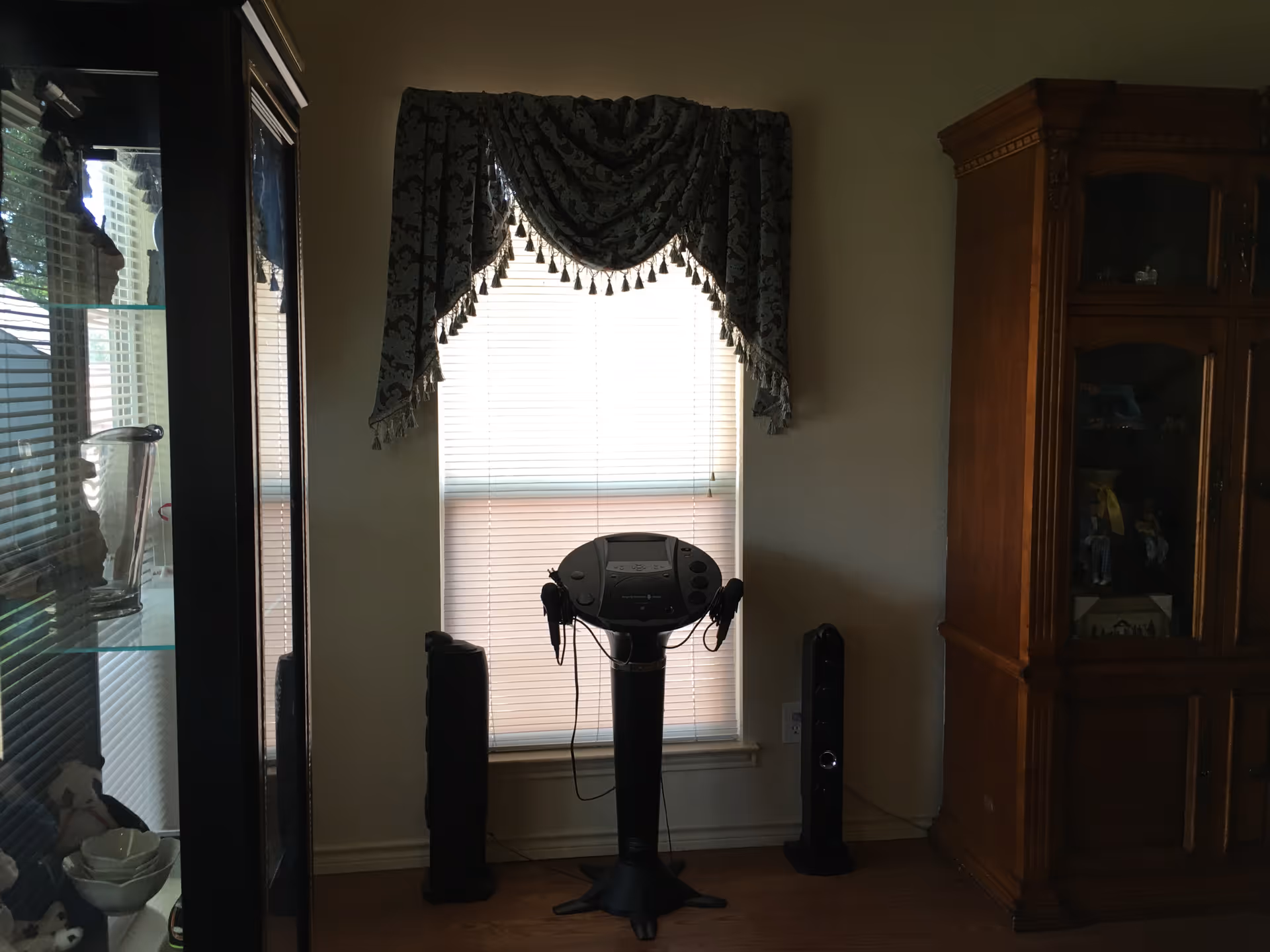 A corner of a room with a window covered by a decorative valance and blinds. In front of the window is a black electronic device on a stand with two attached headphones. To the right is a wooden cabinet with glass doors displaying various items. To the left is a glass display cabinet with shelves holding decorative objects.