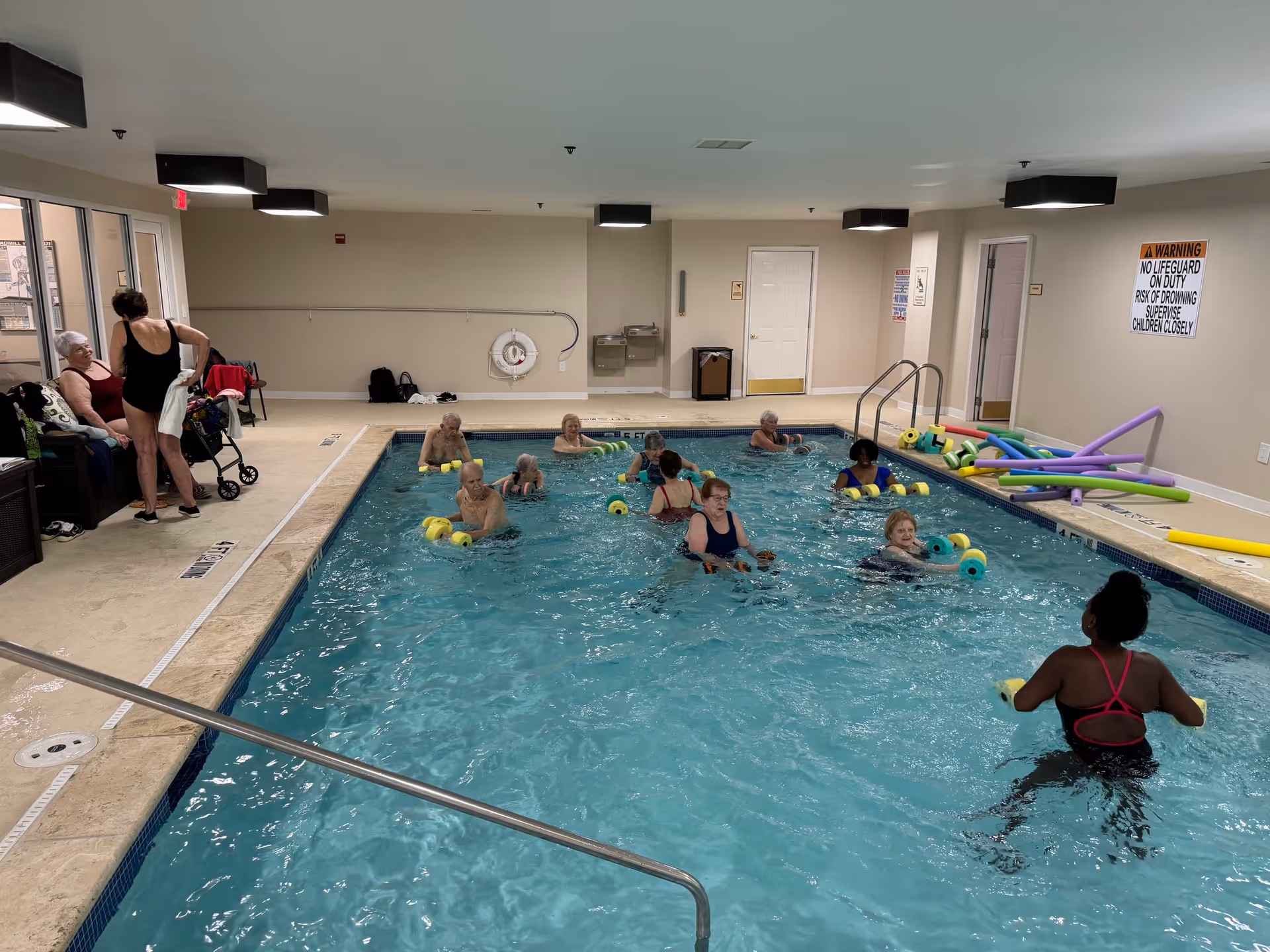 Indoor swimming pool with several elderly people participating in a water exercise class using foam dumbbells. Two women are standing and talking near the poolside, one with a walker. Pool noodles are stacked on the right side near the wall, and a warning sign about no lifeguard on duty is visible on the wall.