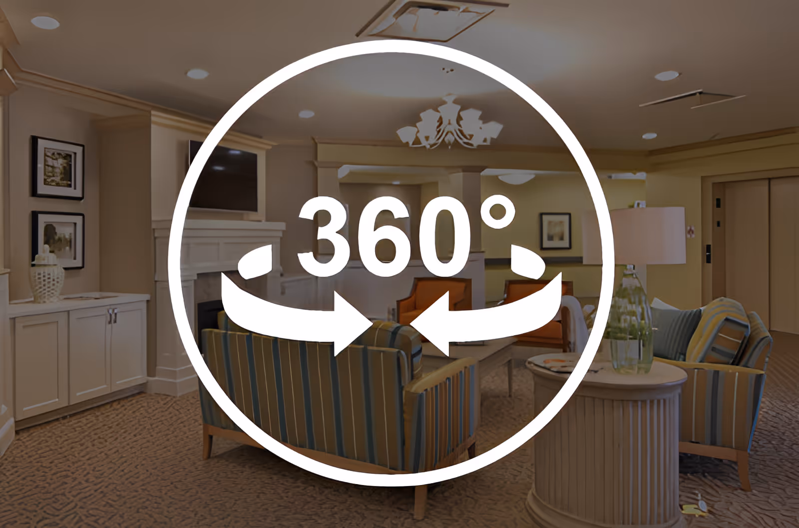 A cozy living room area in a senior living facility with striped armchairs, a coffee table, a fireplace with a mounted TV above it, framed pictures on the wall, and a lamp on a round side table. The room has warm lighting and carpeted flooring.