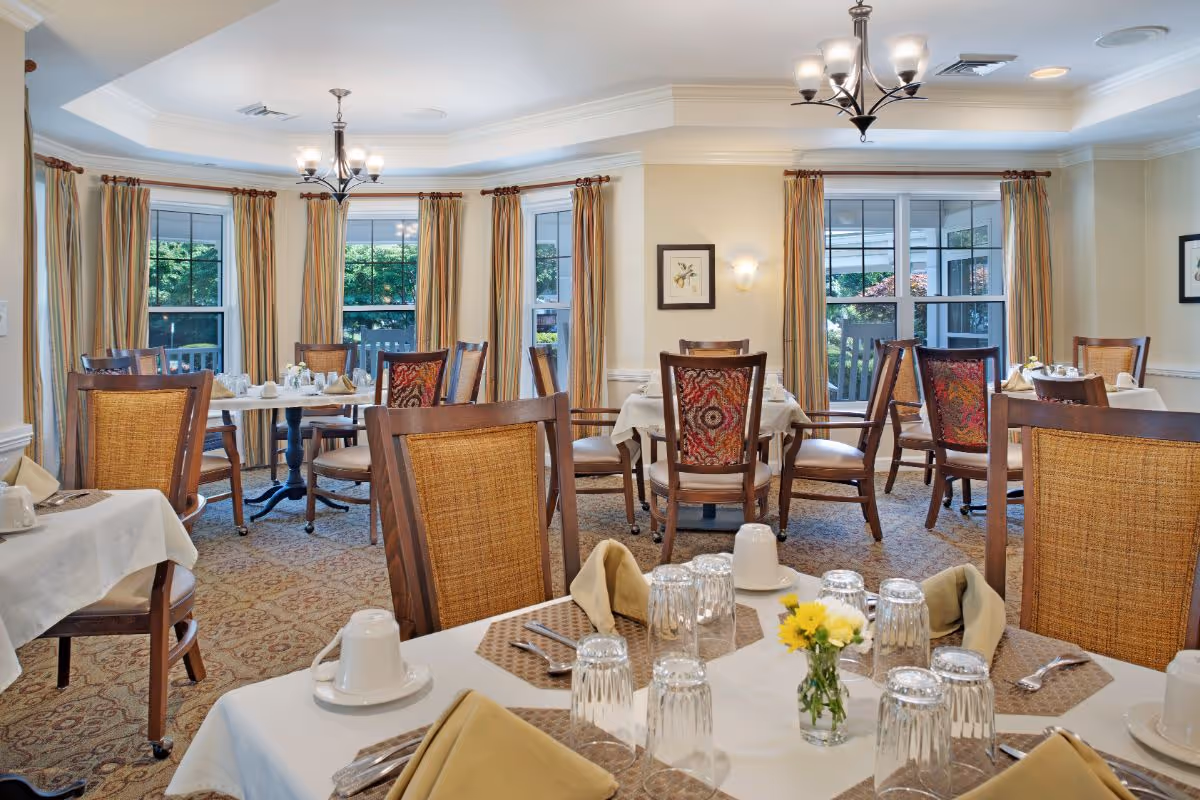 Bright dining room with multiple set tables, wooden chairs, chandeliers, and large windows looking out to greenery.