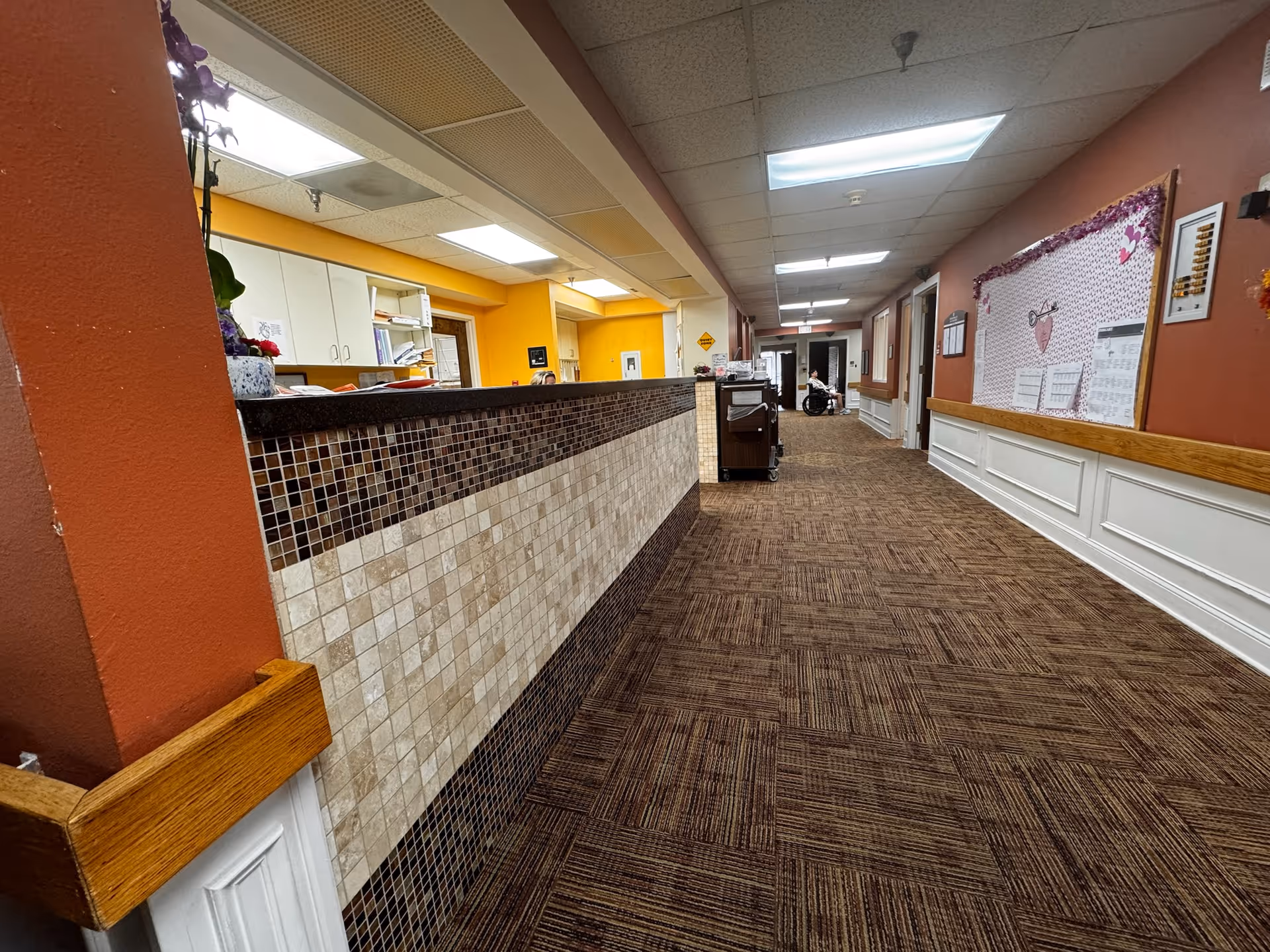 Interior hallway of Heritage Park Health Center with a reception desk on the left side featuring a tiled front. The hallway has brown patterned carpet, orange and white walls, fluorescent ceiling lights, and a bulletin board decorated with pink and purple accents on the right wall. A person in a wheelchair is visible in the distance near the end of the hallway.