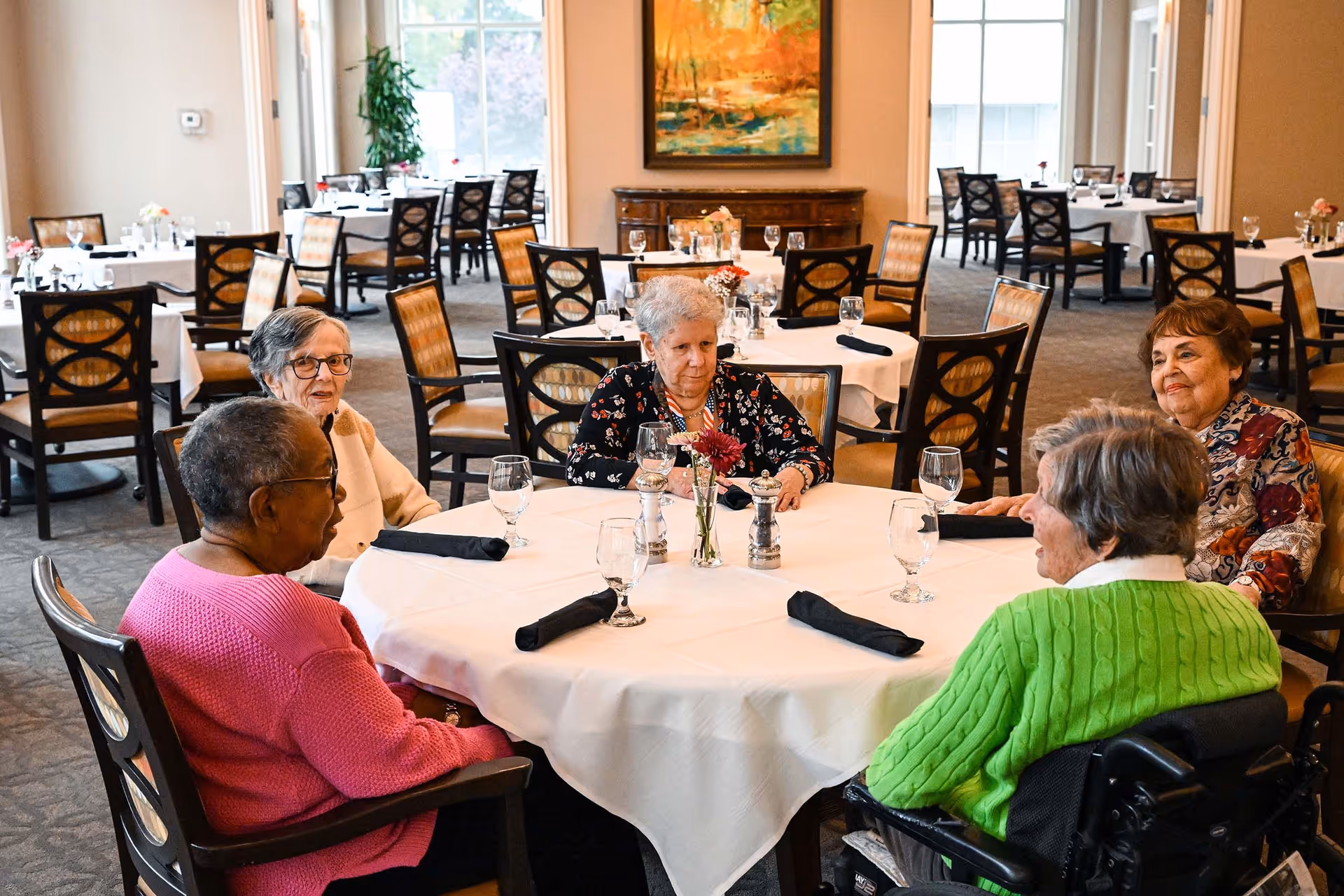 Five elderly women sitting around a round dining table in a well-lit dining room with white tablecloths, black napkins, and glassware. The room has multiple tables and chairs, large windows, and a painting on the wall.