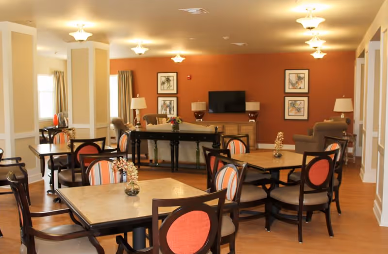 A spacious common area with multiple square tables and chairs featuring orange and beige upholstery. In the background, there is a living room setup with sofas, armchairs, lamps, a TV mounted on an orange accent wall, and framed artwork. The room has warm lighting and wooden flooring.