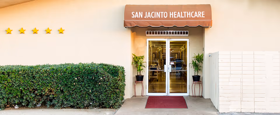 Entrance to San Jacinto Healthcare with double glass doors, a brown awning displaying the facility name, two potted plants on either side of the door, a red doormat, and a hedge to the left with six yellow stars on the wall above it.