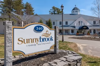 Exterior view of Sunnybrook Senior Living facility with a large white building featuring multiple windows, a cupola, and a covered entrance. In the foreground, there is a stone-bordered sign displaying the name 'Sunnybrook' and the address number 340. Trees and a clear sky are visible in the background.