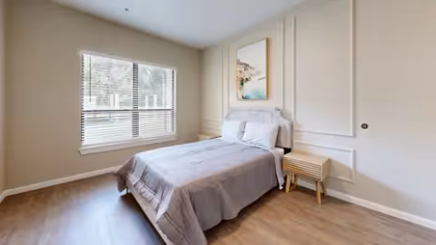 A simple bedroom with a neatly made bed covered with a gray comforter and white pillows. There is a wooden nightstand with a drawer next to the bed. A large window with white blinds allows natural light to enter the room. The walls are painted beige with decorative molding, and a framed abstract artwork hangs above the bed. The floor is wooden.