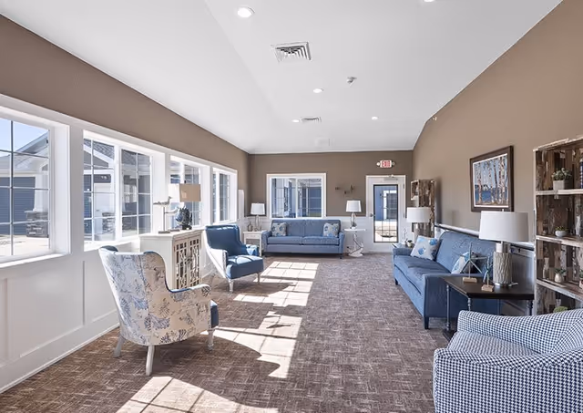 A bright and spacious living room area with large windows on the left side letting in natural light. The room features multiple blue upholstered sofas and armchairs arranged along the walls, with side tables and lamps. The walls are painted brown, and there is a carpeted floor. Decorative shelves and framed artwork are visible on the right wall.