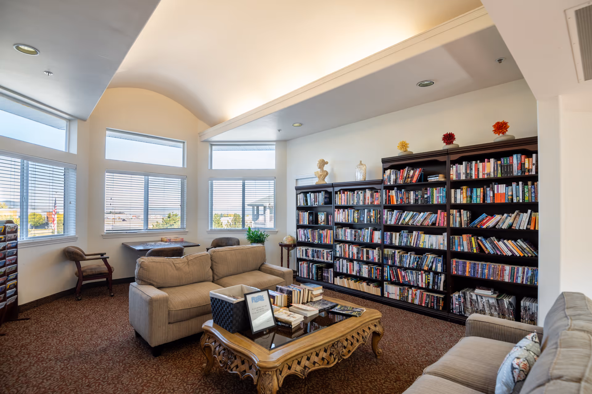 A cozy senior living facility library room with large windows letting in natural light. The room features two beige sofas facing a wooden coffee table with books and a basket on it. Along one wall, there are tall dark wood bookshelves filled with books and decorative items on top. There are also a couple of chairs and a small table near the windows.