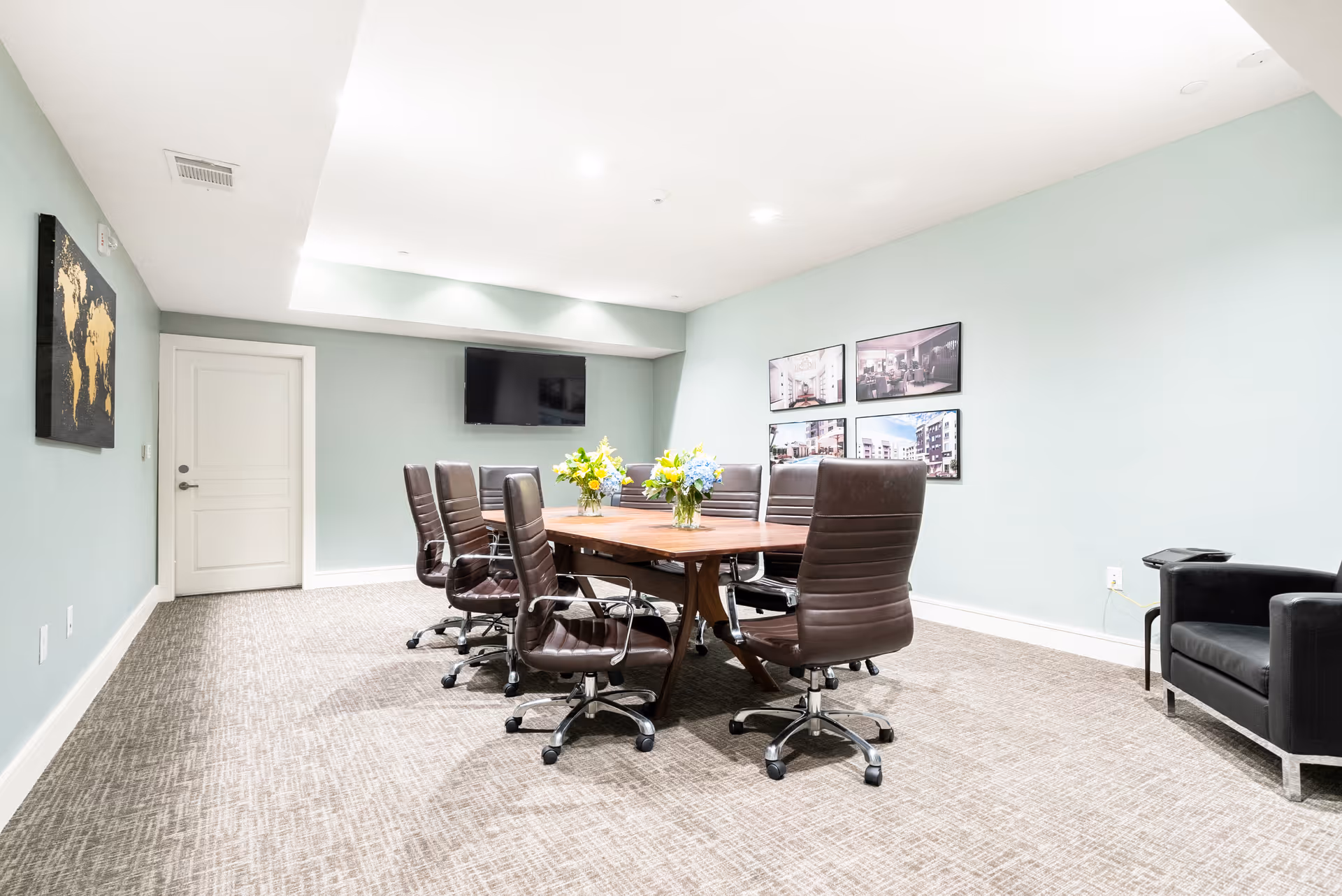 A modern conference room with a wooden table surrounded by eight brown leather swivel chairs. The room has light blue walls, a beige carpet, a flat-screen TV mounted on the wall, and framed pictures on one wall. There is a black armchair in the corner and two vases with flowers on the table.