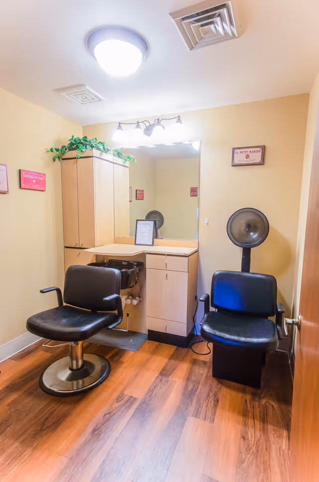 A small salon room with two black salon chairs, a wooden cabinet with a countertop and a large mirror above it. There is a hair dryer hood attached to the wall next to one of the chairs. The room has wooden flooring and beige walls with small framed pictures and a green plant on top of the cabinet.
