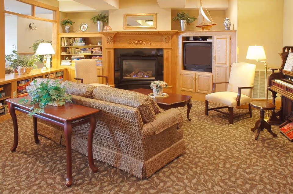 A cozy living room area with patterned carpet and a matching patterned sofa facing a wooden fireplace with a small fire burning. The fireplace is flanked by built-in wooden shelves filled with books and board games on the left, and a TV on the right. Two upholstered armchairs with wooden arms are positioned near the fireplace, accompanied by side tables and lamps. A wooden table behind the sofa holds a green potted plant. A piano is visible on the right side of the room.