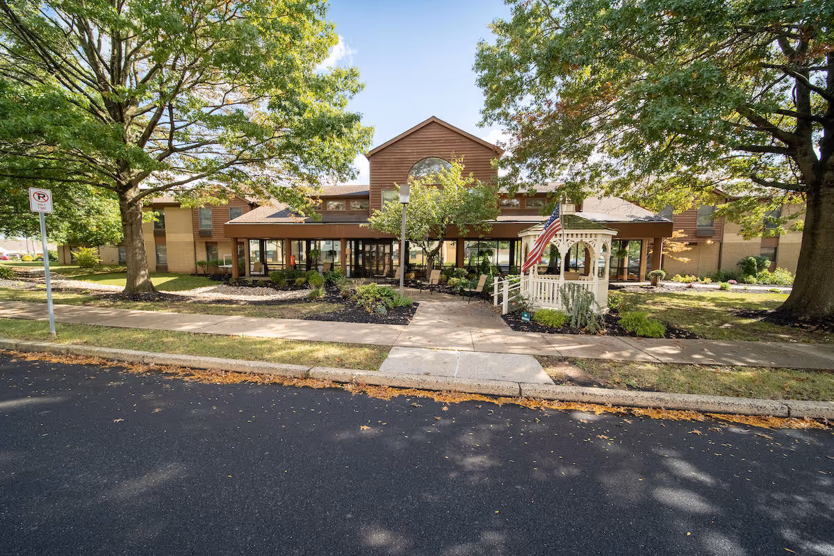 Front exterior view of Independence Court of Quakertown building with a pathway leading to the entrance, surrounded by trees and landscaping including a small white gazebo with an American flag.