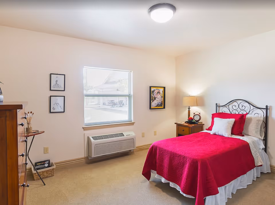 A cozy bedroom with a single bed covered in a bright red quilt and white pillows. The bed has a decorative black metal headboard. Next to the bed is a wooden nightstand with a lamp, a clock, and some small decorative items. On the wall above the nightstand is a framed painting. There is a window with blinds letting in natural light, and two small framed sketches hang on the adjacent wall. A wooden dresser and a small round table with art supplies are also visible. The room has beige carpet and light-colored walls.