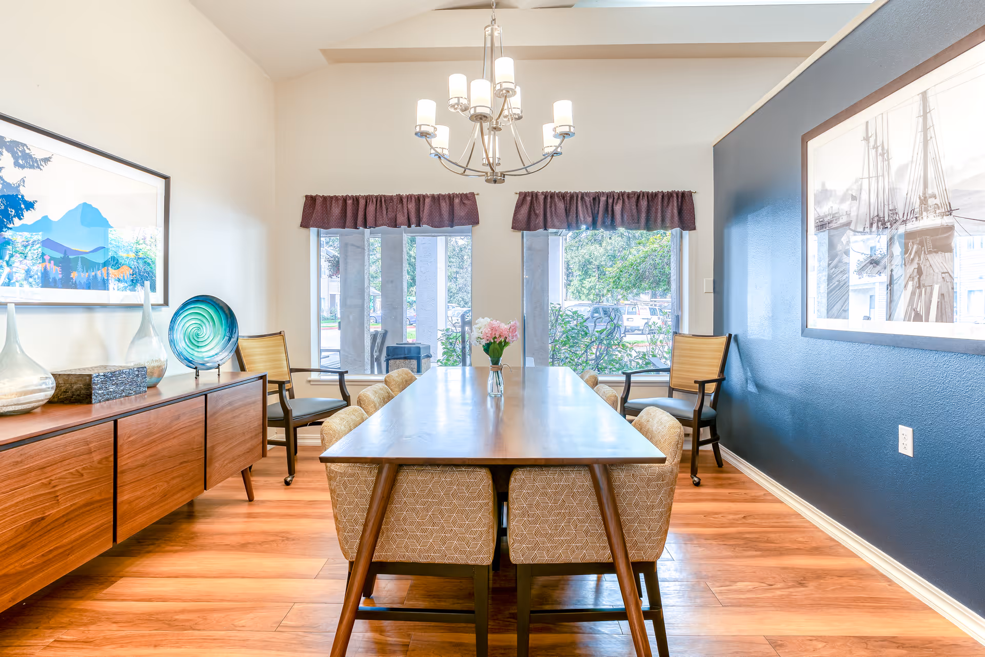 A bright dining room with a wooden table surrounded by eight upholstered chairs. The room features a wooden sideboard with decorative glass vases and a colorful plate. Two large windows with purple valances allow natural light to enter, and a modern chandelier hangs above the table. The walls are painted white and dark blue, with framed artwork on each side.