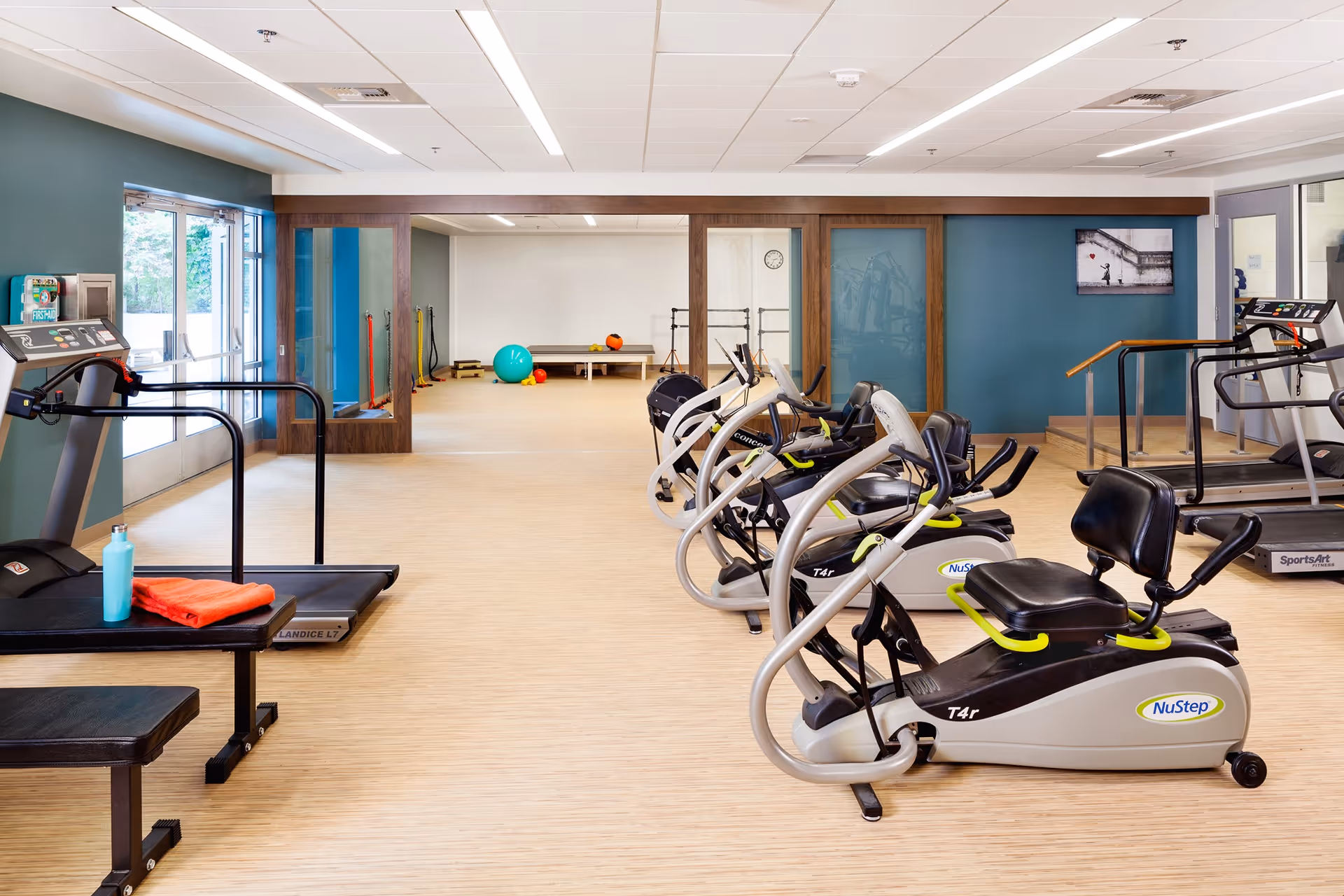 A bright and spacious fitness room in Bayview Retirement Community featuring several exercise machines including recumbent bikes and treadmills. The room has light wood flooring, teal walls, and large windows letting in natural light. In the background, there is an open area with exercise balls and other fitness equipment.