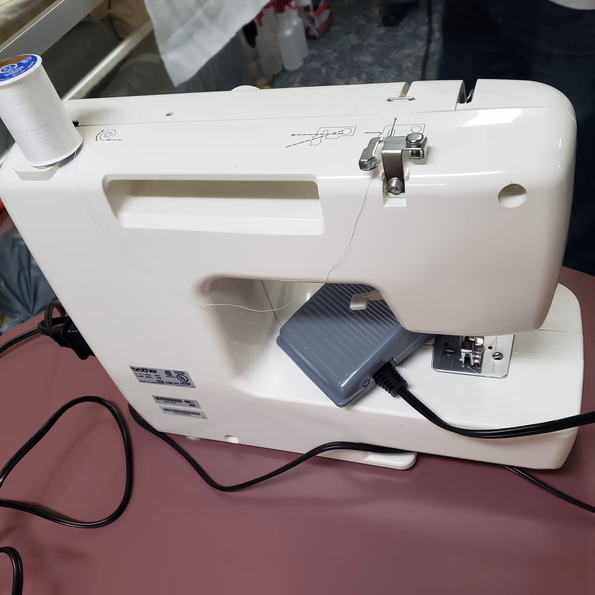 A white Brother sewing machine placed on a maroon surface with a spool of white thread on top and a foot pedal connected by a black cord.