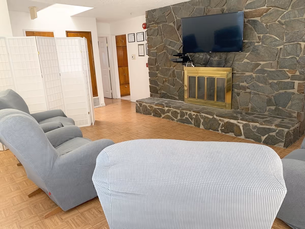 A cozy living room area with three light gray upholstered armchairs facing a stone fireplace with a brass frame. Above the fireplace is a mounted flat-screen TV. The room has a wooden floor and white walls, with a white folding room divider and wooden doors visible in the background.