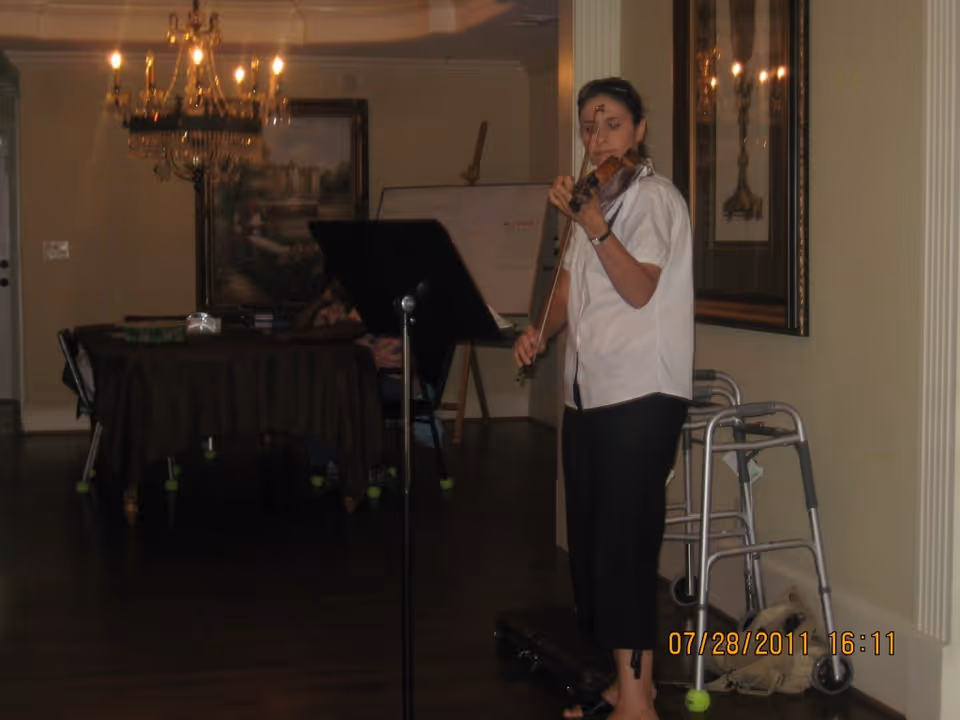 A woman playing the violin indoors near a walker. Behind her is a music stand, a table with a dark tablecloth, chairs, a chandelier, and framed artwork on the walls.