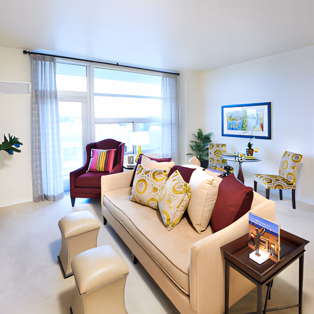 Bright modern living room with a beige sofa, colorful cushions, a burgundy armchair, and a large window with sheer curtains.