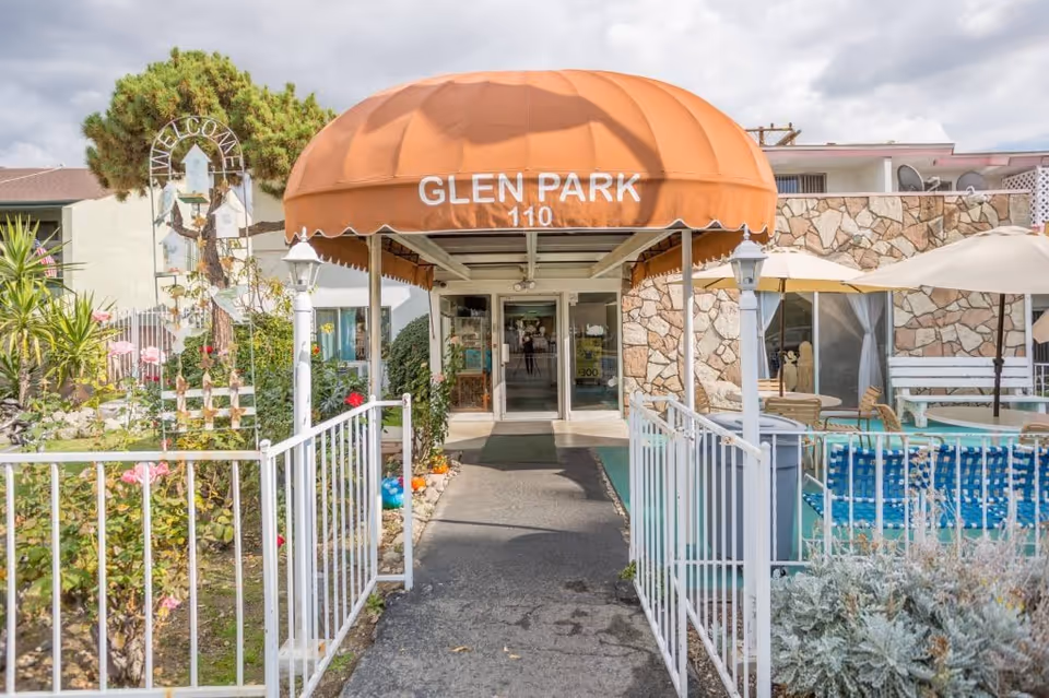 Entrance to Glen Park at Monrovia facility with an orange canopy displaying the name and address number 110. The pathway is flanked by white railings and garden areas with flowers on the left and outdoor seating with umbrellas on the right. The building exterior features stone and white walls under a cloudy sky.