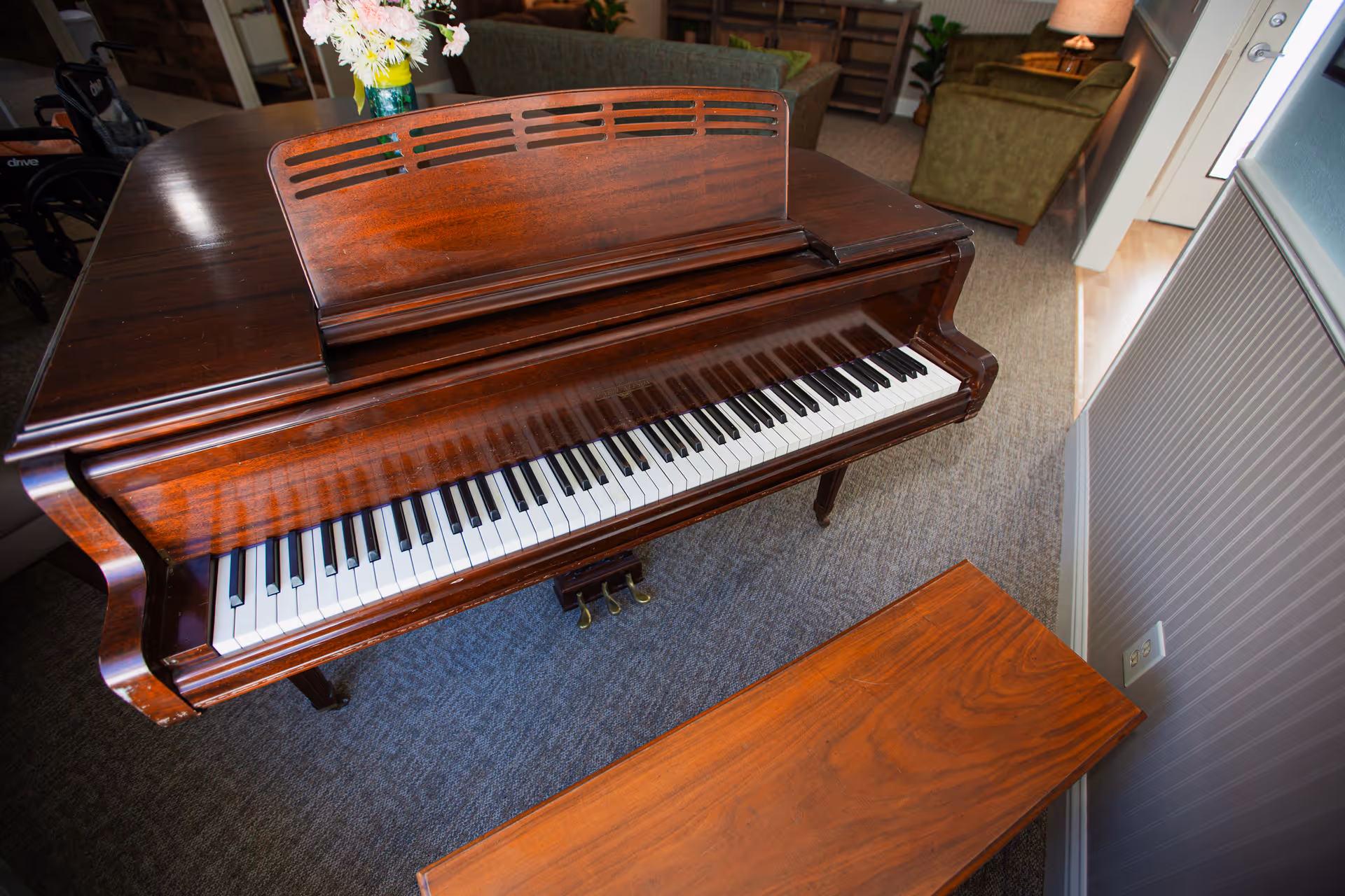 A polished wooden grand piano with an open keyboard cover, placed on a carpeted floor in a room with green upholstered chairs and a vase of flowers on top of the piano.