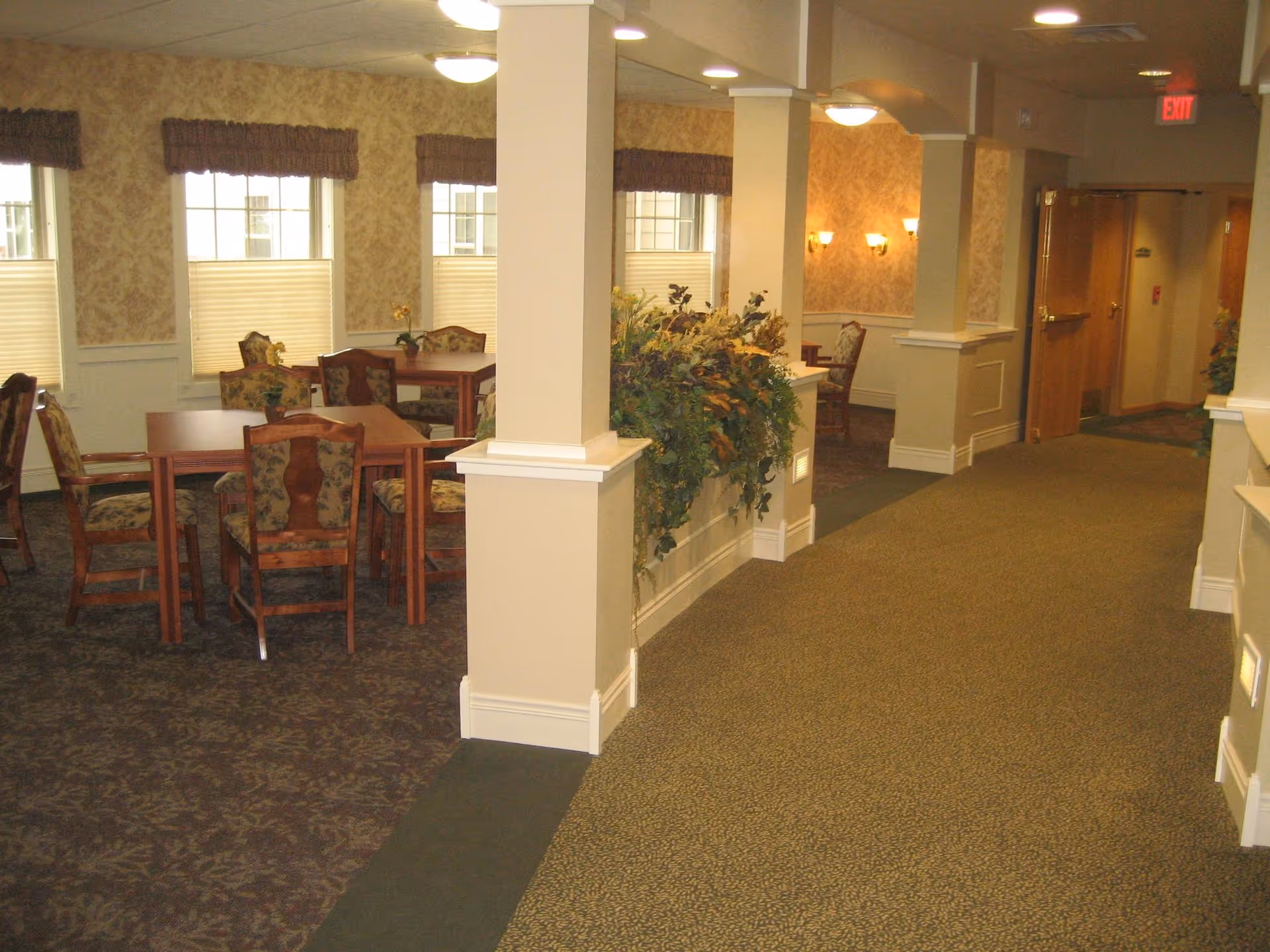 Interior view of a retirement community common area with carpeted floors, beige walls, and multiple wooden tables and chairs with floral upholstery. There are several windows with blinds and valances, decorative plants on a half wall, and ceiling lights illuminating the space. A hallway with an exit sign is visible in the background.