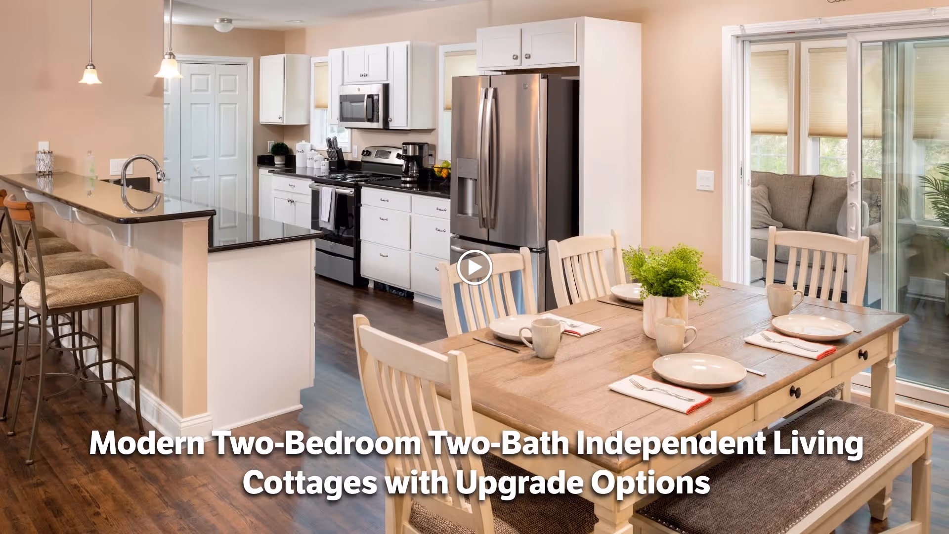 Modern kitchen and dining area in an independent living cottage. The kitchen features white cabinets, stainless steel appliances including a refrigerator, stove, and microwave. There is a breakfast bar with three cushioned stools. The dining table is set with plates, mugs, and napkins, and has a small green plant centerpiece. Sliding glass doors lead to a sunroom with a sofa and plants.