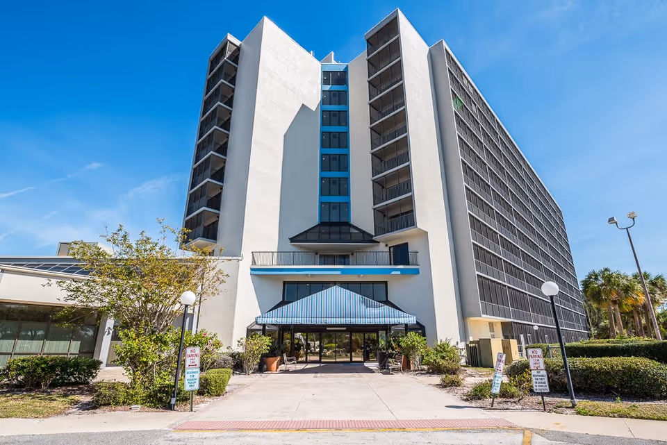 Front entrance and facade of a multi-story senior living building with a covered entryway and balconies under a clear blue sky.