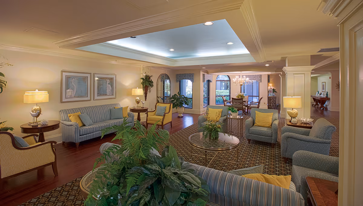 A spacious and well-lit living room area in Regency Oaks Health Center featuring multiple upholstered sofas and armchairs with yellow and blue cushions, a patterned carpet, several side tables with lamps, framed bird artwork on the wall, and a dining area with a chandelier visible in the background. There are large windows allowing natural light and several green plants adding a touch of nature to the room.