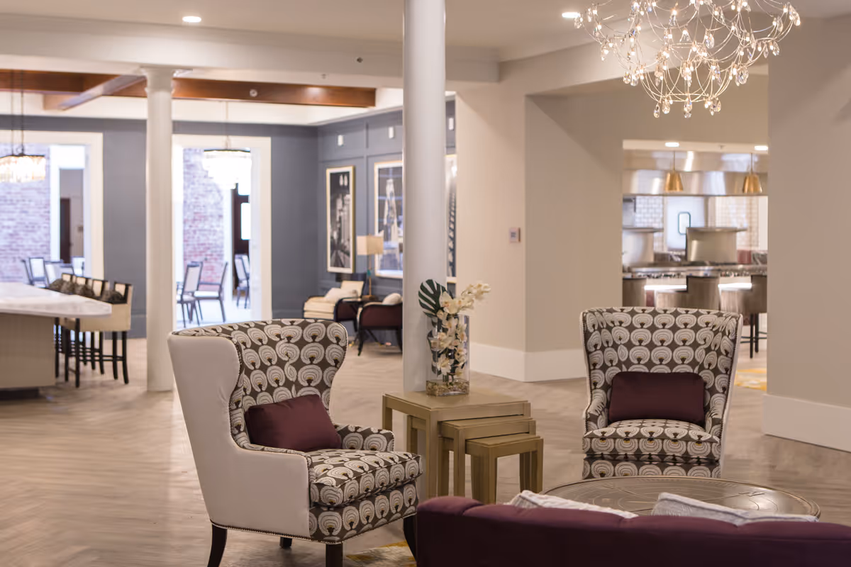 A stylish and spacious interior common area with patterned armchairs featuring purple cushions, a round coffee table, and a set of nested side tables with a vase of flowers. In the background, there is a dining area with chairs and tables, and a kitchen area with bar stools and pendant lights. The space has light-colored walls, wooden flooring, and modern chandeliers.