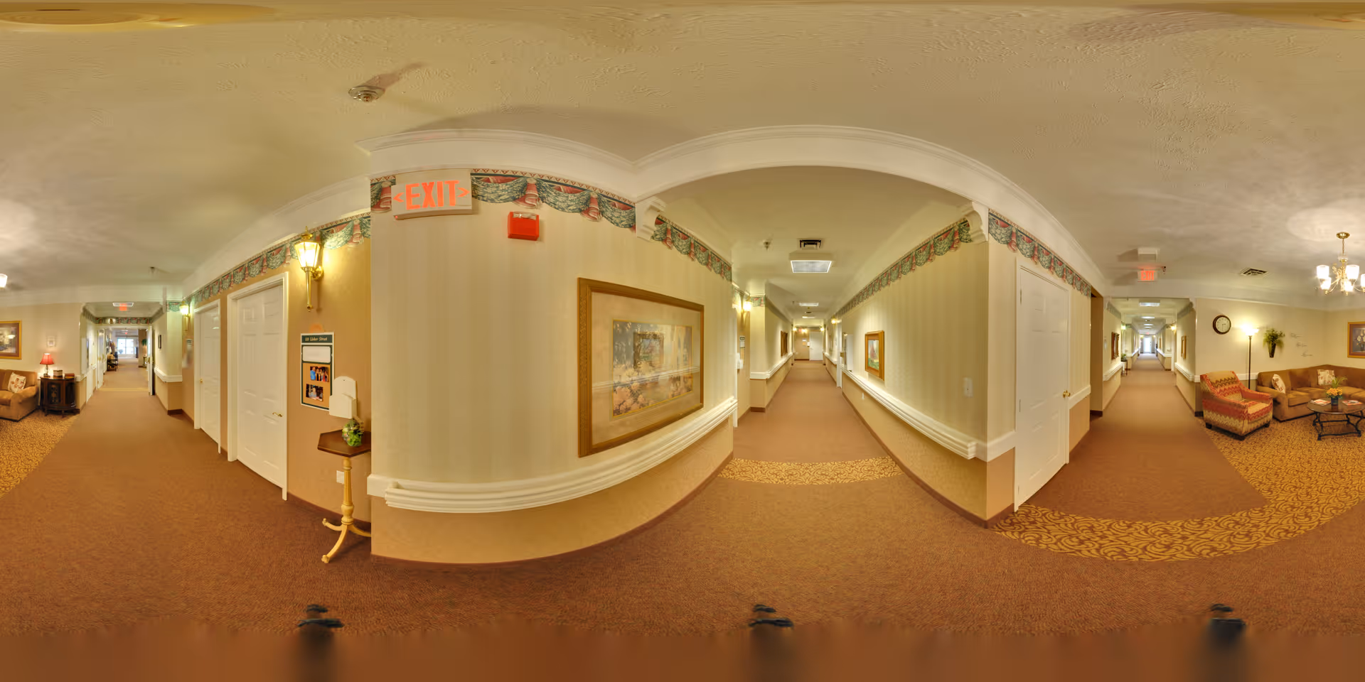 Panoramic carpeted hallway of a senior living facility with seating areas, framed artwork, and exit signs.