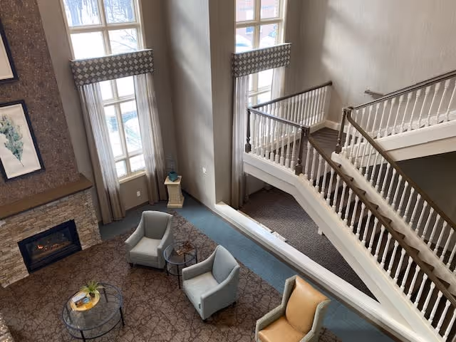 View from above of a cozy living area with a stone fireplace, two gray armchairs, a tan armchair, a round glass coffee table with a plant and books, large windows with curtains, and a staircase with white railings.