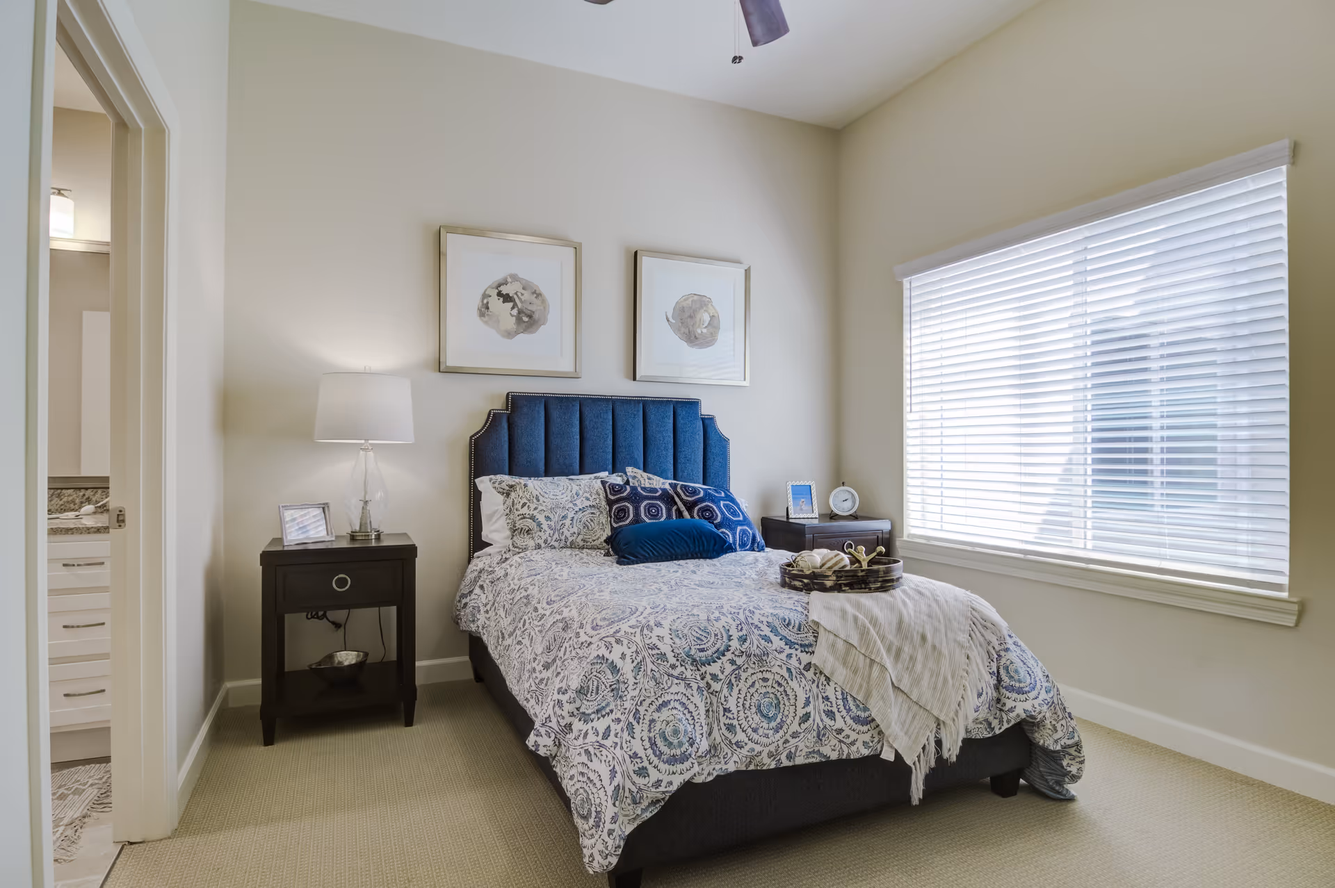Sunlit bedroom with a patterned bed, blue upholstered headboard, nightstands with a lamp, and a large window with blinds.