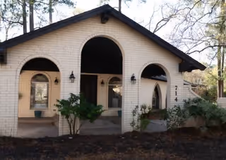 Exterior view of a single-story brick building with three arched openings on the front porch. The building has a dark roof and is surrounded by trees and some shrubbery. The number 714 is visible on the right side of the building.
