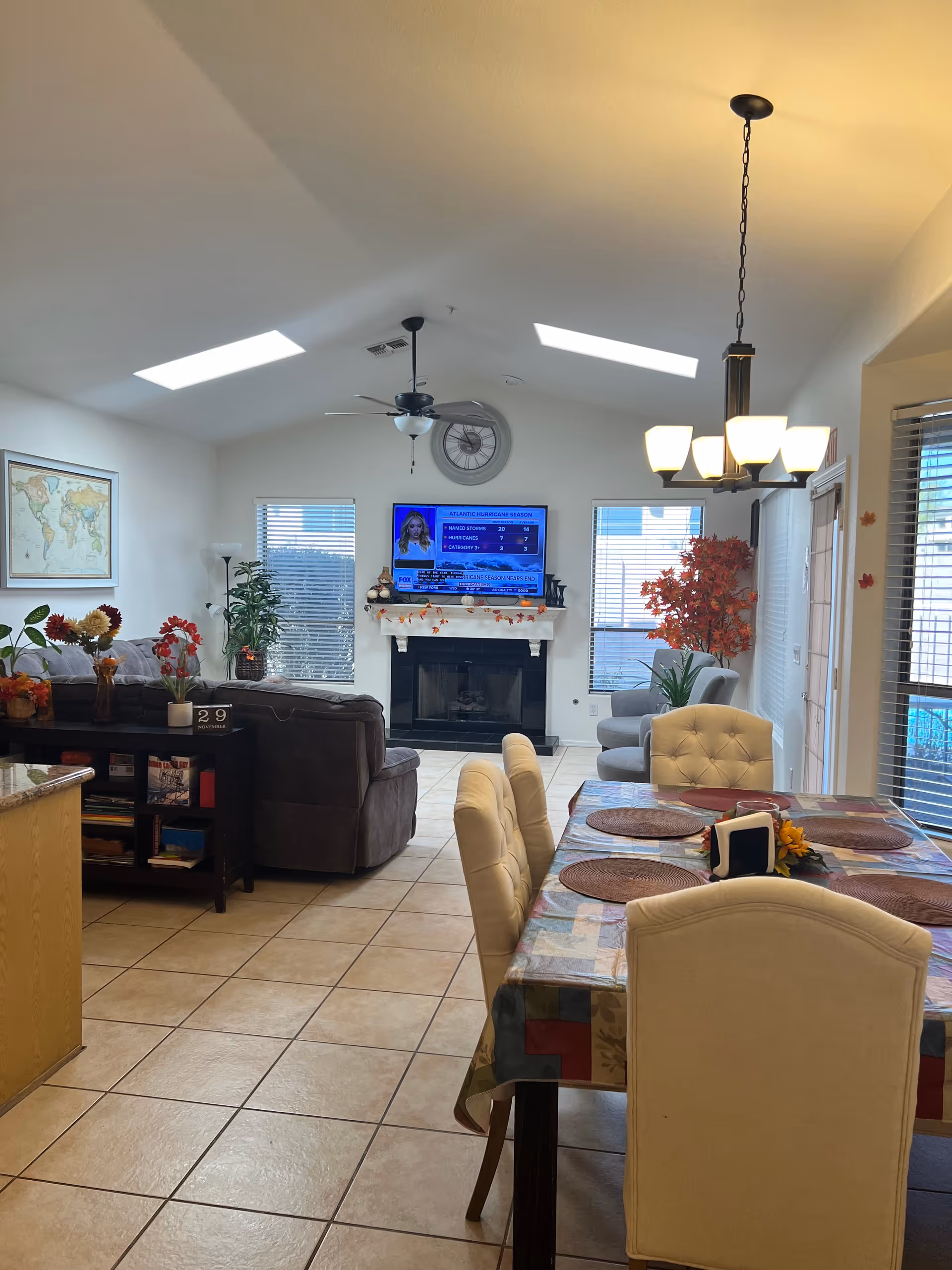 A bright and cozy living and dining area with tiled floors. The dining table is covered with a patterned tablecloth and set with placemats and a small centerpiece. The living room features a dark brown sectional sofa, a fireplace with seasonal decorations, a wall-mounted TV showing a news channel, and large windows with blinds. There are ceiling fans and a hanging light fixture, along with decorative plants and a world map on the wall.