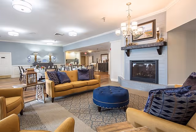 A spacious and well-lit living room area in TerraBella Athens featuring mustard yellow sofas with dark blue cushions, a round blue ottoman, a fireplace with a wooden mantel displaying decorative items and a framed picture, and a dining area with tables and chairs in the background.