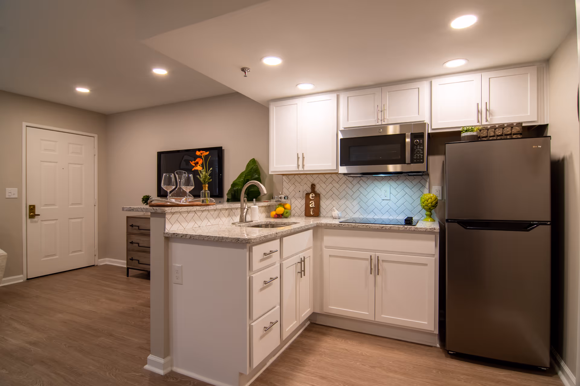 Modern kitchen area in a retirement community featuring white cabinets, a stainless steel refrigerator and microwave, a small sink with a curved faucet, and a countertop with decorative items including a vase with orange flowers, two wine glasses, and a bowl of fruit. The kitchen has a light-colored herringbone tile backsplash and wood flooring. In the background, there is a white door and a small dresser with a plant on top.