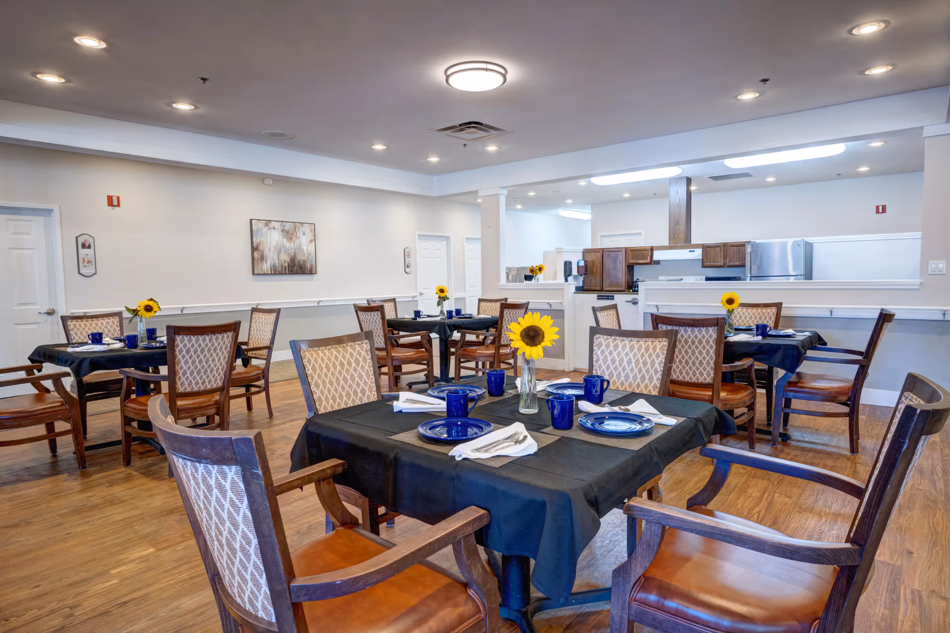 A dining room in a senior living facility with several tables covered in black tablecloths, each set with blue plates, blue mugs, silverware, and white napkins. Each table has a vase with a sunflower. The room has wooden floors, beige walls, recessed lighting, and an open kitchen area with wooden cabinets and stainless steel appliances in the background.