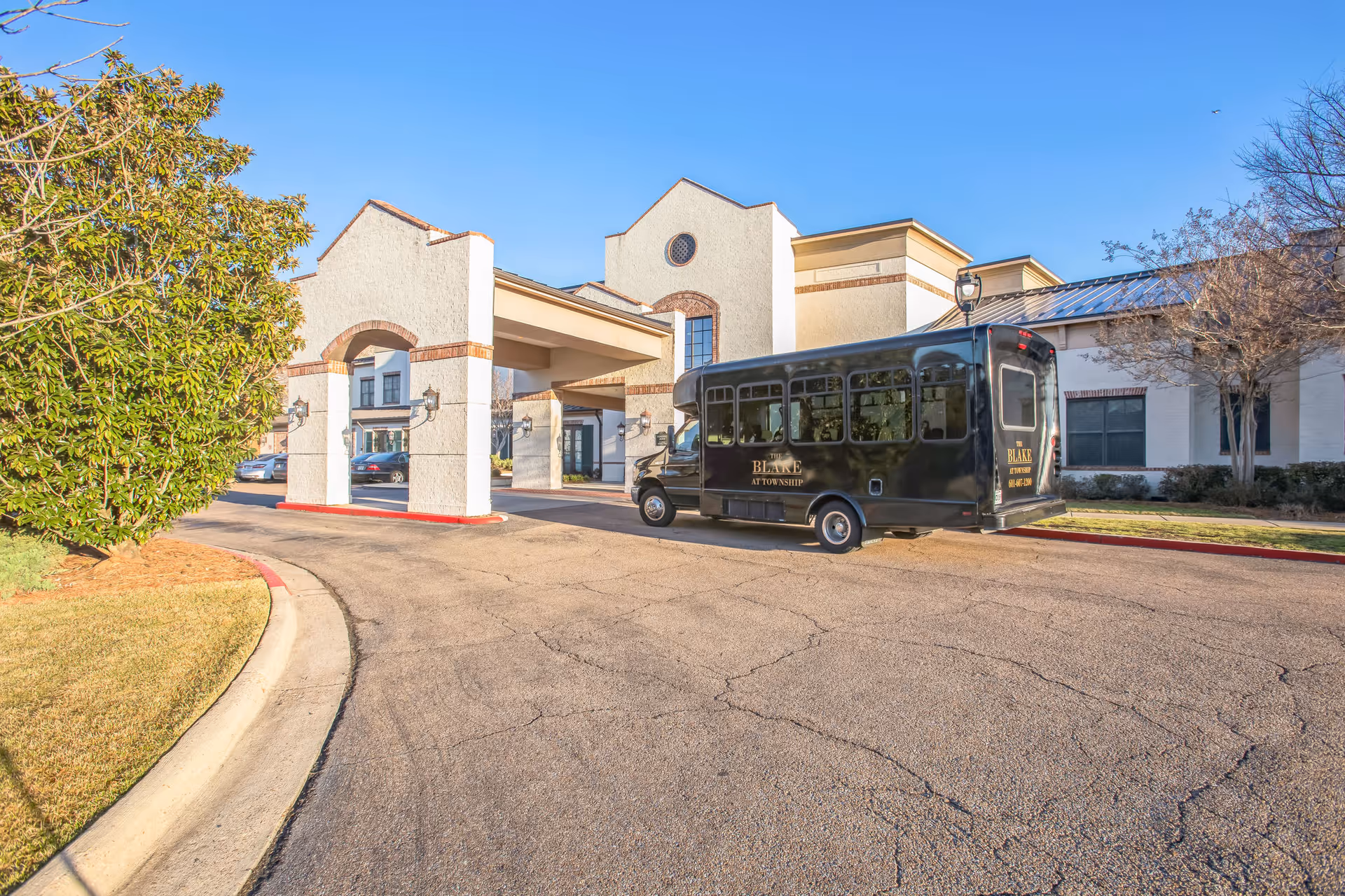 Exterior view of The Blake at Township senior living facility showing the entrance with a covered drop-off area and a black shuttle bus parked nearby. The building has a light-colored facade with brick accents and is surrounded by trees and landscaping under a clear blue sky.