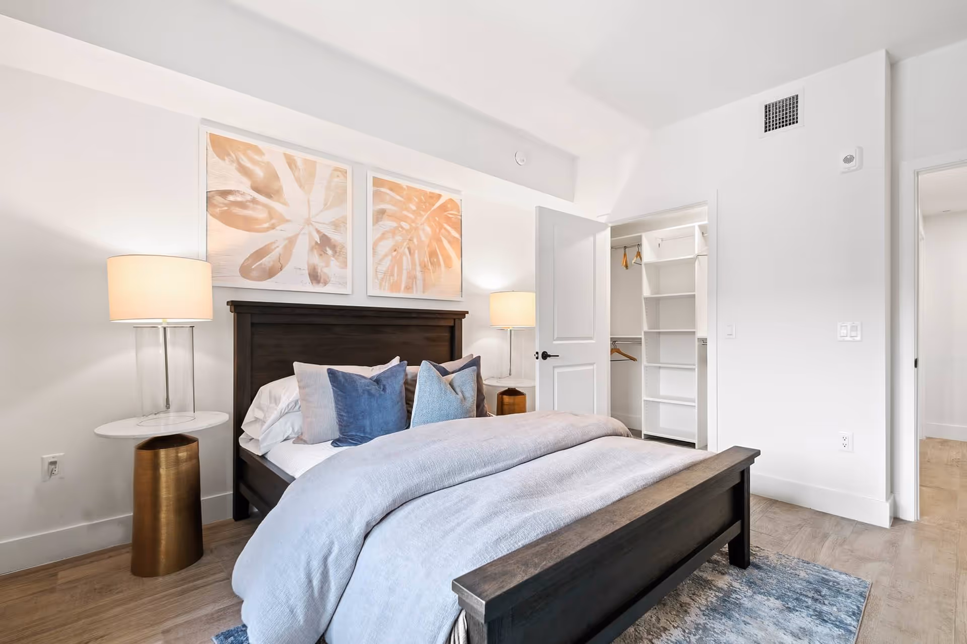 Bright modern bedroom with a dark wood bed, gray bedding, bedside lamps, artwork above the headboard, and an open walk-in closet.