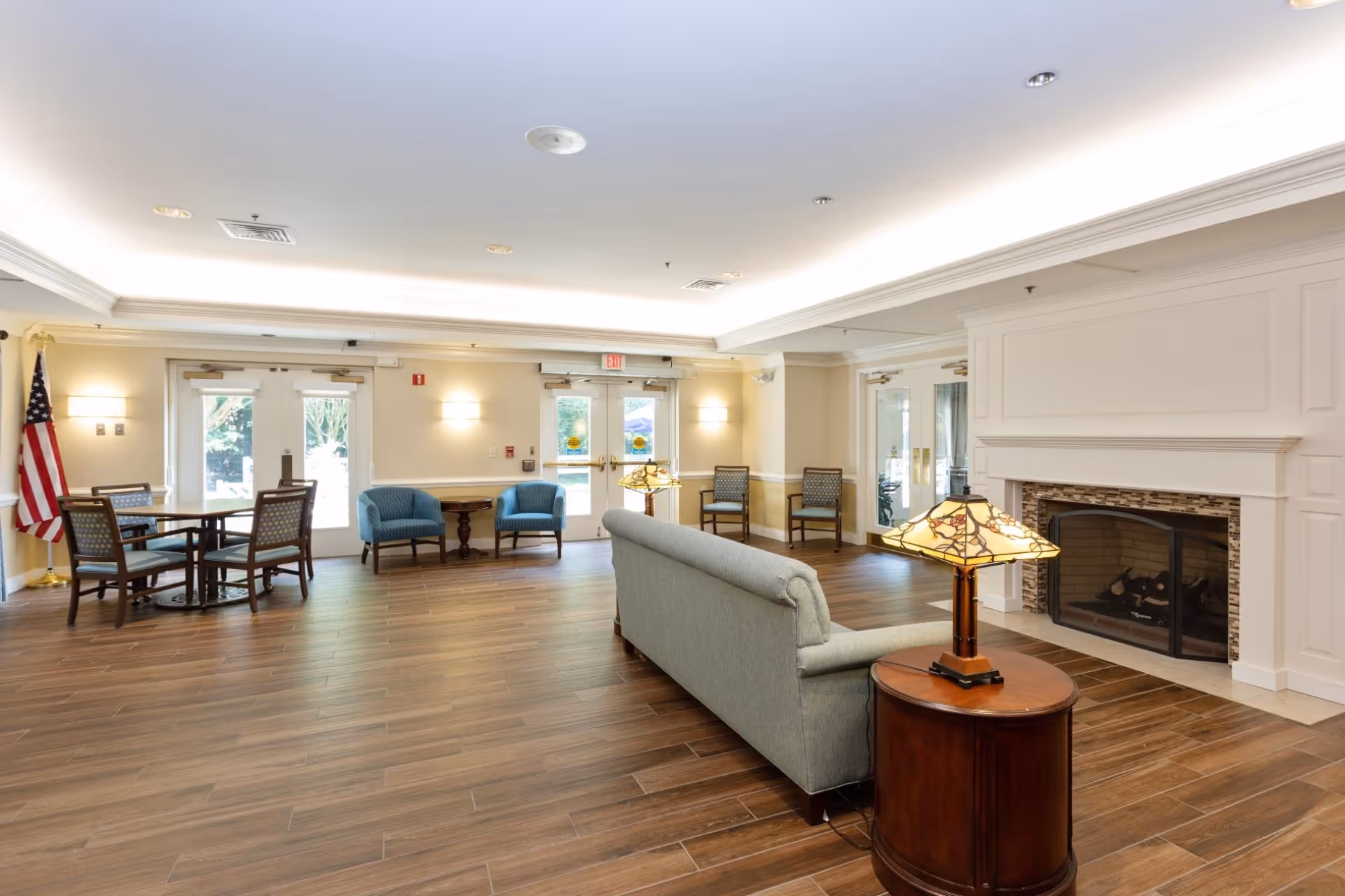 Bright communal lounge with a sofa, armchairs, tables, a fireplace, and wood-look flooring.